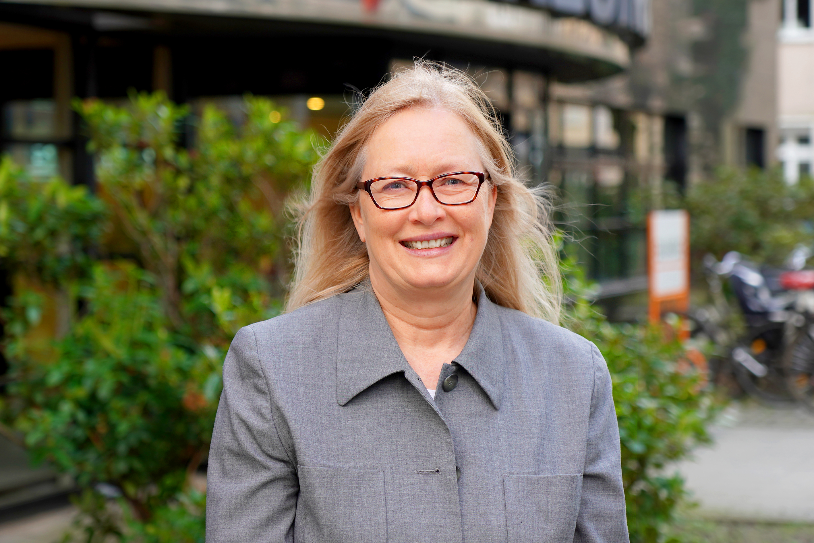You can see a woman with blonde hair and black glasses. She is also wearing a grey blazer and there are bushes in the background.