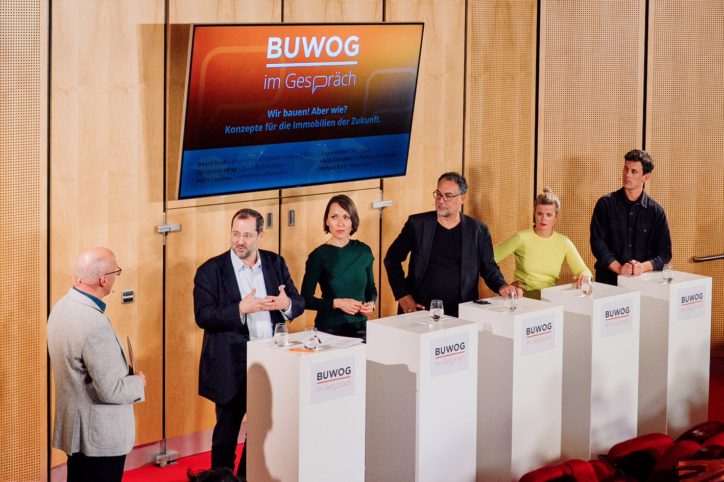 You can see 6 people standing on a stage at desks discussing a topic.