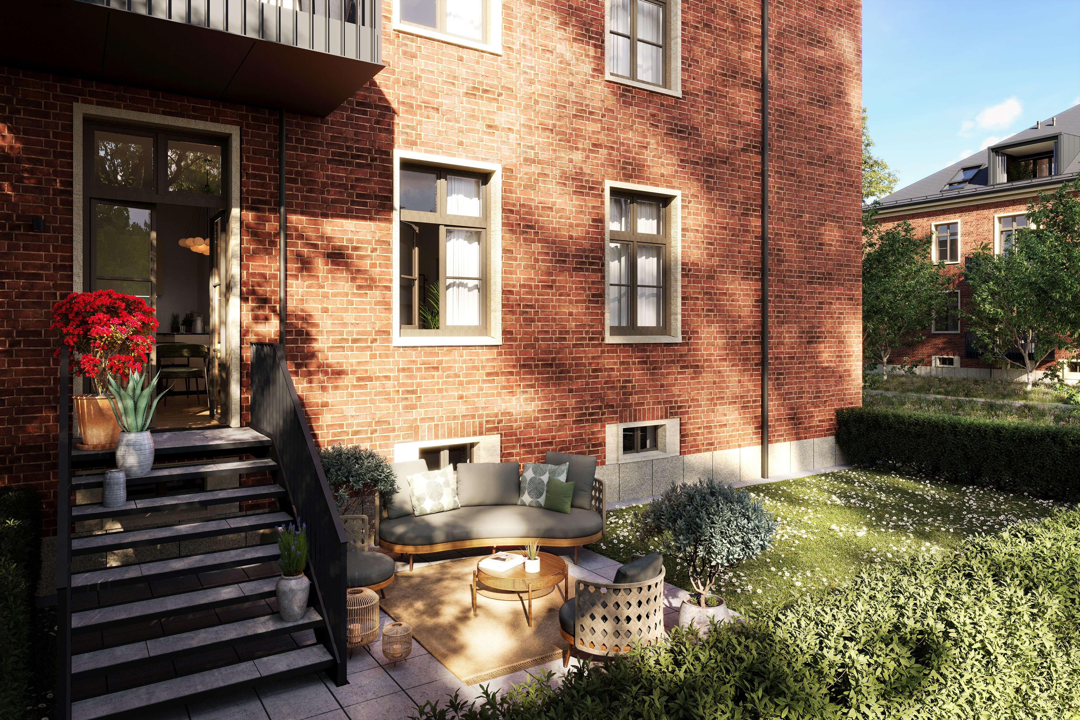 Zu sehen ist eine Terrasse im Erdgeschoss mit Treppen zum Wohnungseingang. Auf der Terrasse ist eine Couch mit Teppich, Couchtisch und Stühlen. Auf den Treppen stehen Blumen.