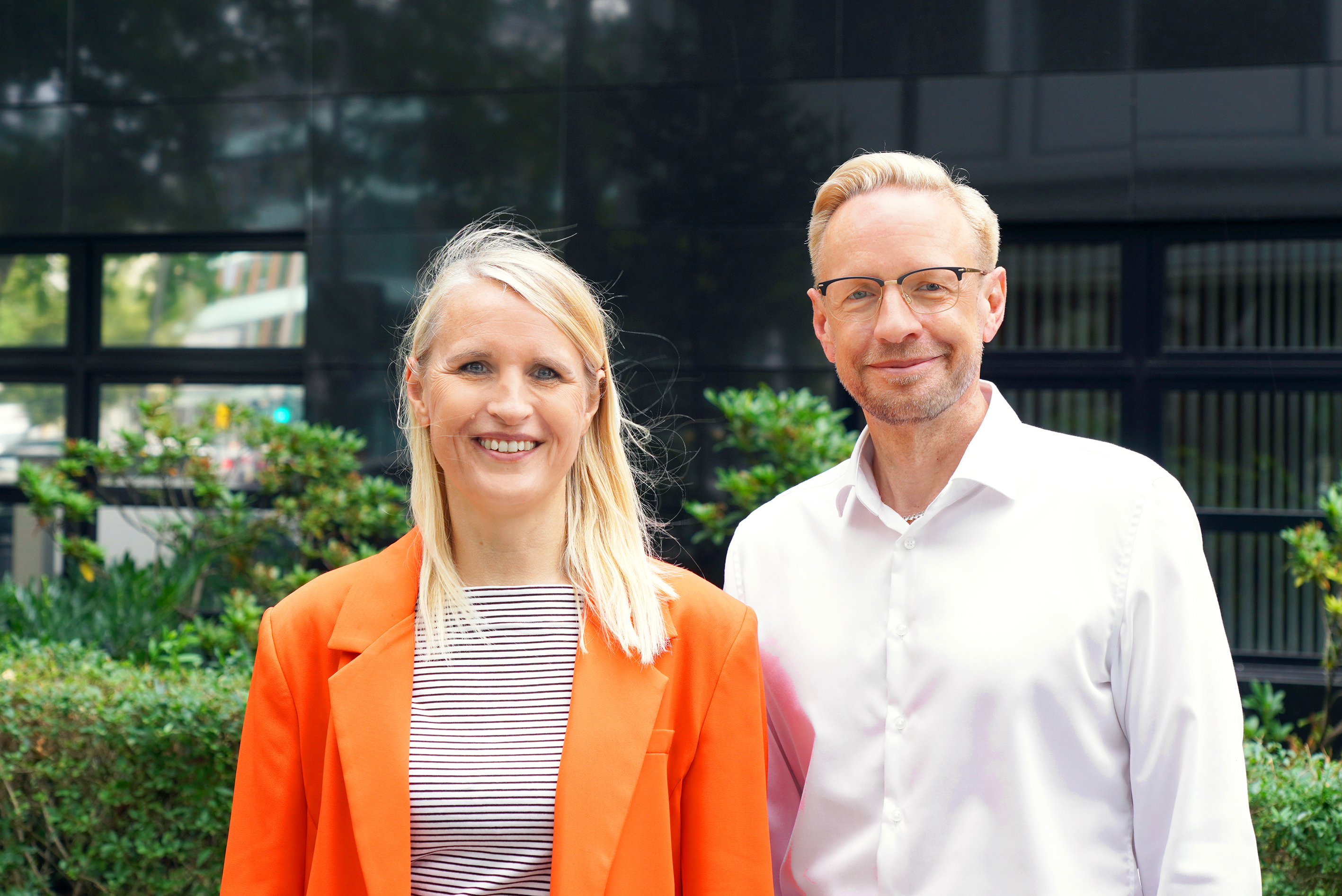 On the left is a woman with blonde hair and an orange blazer, and on the right is a man with short blonde hair and a white shirt. In the background, you can see greenery and windows.
