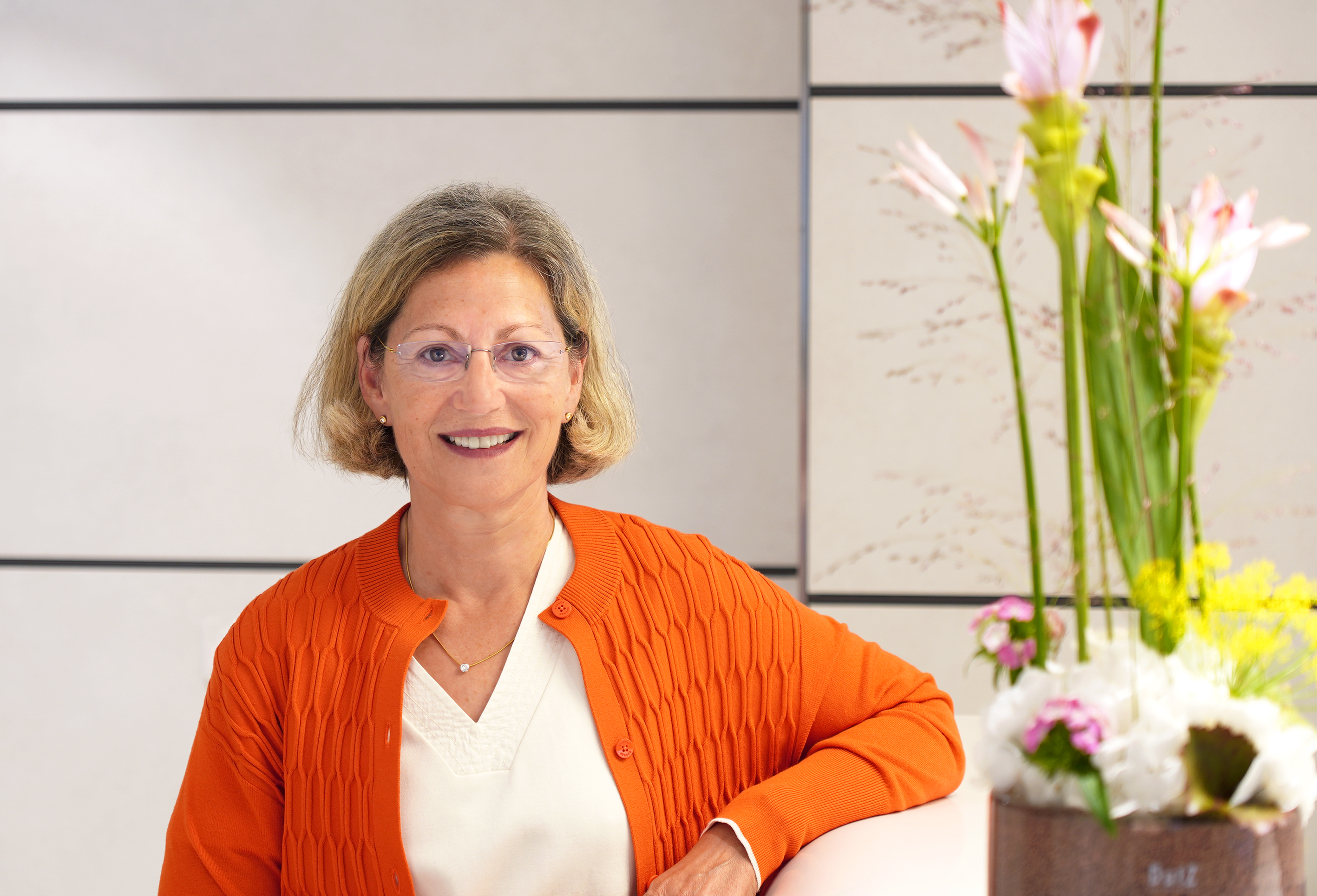 The picture shows a woman with chin-length blonde hair wearing an orange cardigan. There are flowers in the foreground and the background is white. She is leaning her left arm on a counter.