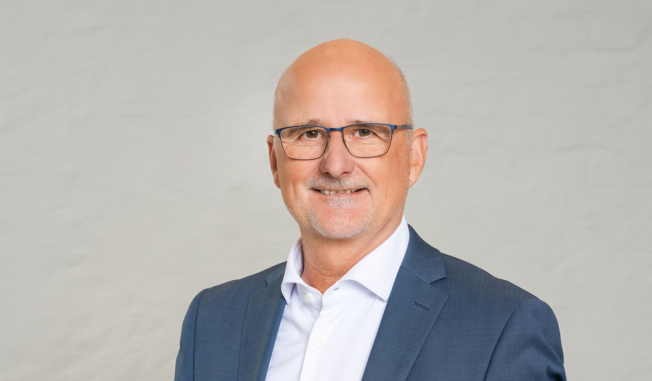 You can see a man in a white shirt and a blue blazer. He is bald and wears large blue glasses.